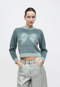 Τυρκουάζ cropped πουλόβερ με υφή πλέξης και μοτίβο φιόγκου. Συνδυάζεται με ασημί μεταλλικό παντελόνι φαρδιάς γραμμής.