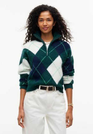 Mujer con pelo rizado que lleva un suéter de rombos verde, azul marino y blanco, pantalones blancos de pernera ancha, cinturón negro y mocasines marrón oscuro.