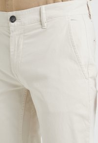 Béžové bavlněné kalhoty se slim fit střihem, modrým knoflíkem a bočními kapsami, jemnou texturou a jednoduchým designem.