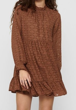 Kobieta w brązowej, długorękawowej mini sukience z fakturą, z falbaną na dole i wysokim dekoltem, stojąca na tle gładkiej ściany.