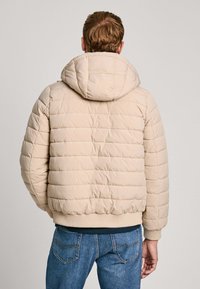 Chaqueta de plumas beige con capucha, con diseño acolchado y puños elásticos, combinada con pantalones de mezclilla de corte relajado.