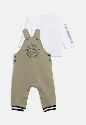Salopette bébé beige avec logo noir et boutons-pression, associée à un t-shirt blanc à manches longues avec texte du logo sur une manche.