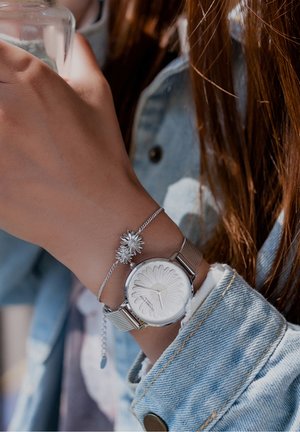 Poignet avec montre à bracelet en maille argentée ornée d'un motif floral et un bracelet assorti en forme de fleur, portant une veste en denim bleu clair.