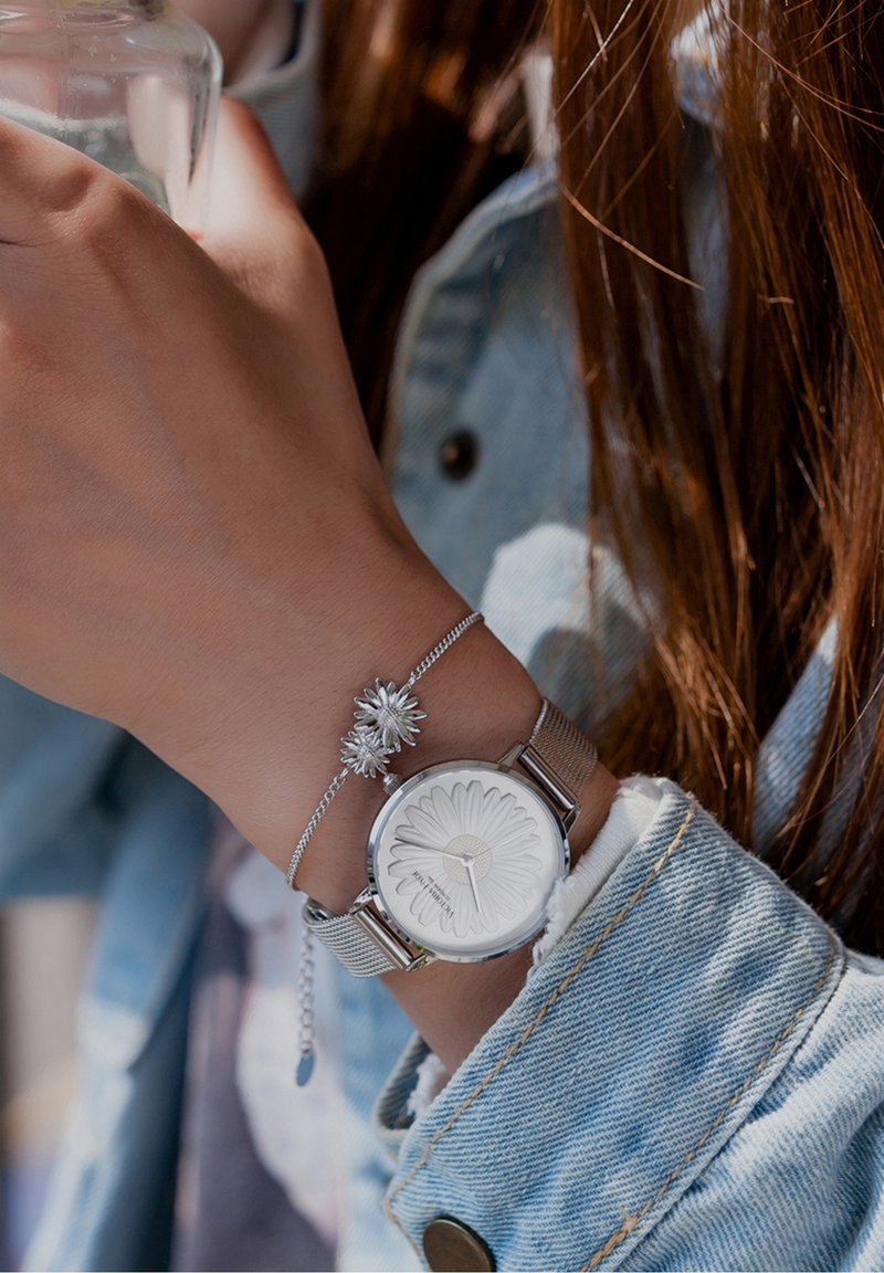 Poignet avec montre à bracelet en maille argentée ornée d'un motif floral et un bracelet assorti en forme de fleur, portant une veste en denim bleu clair.