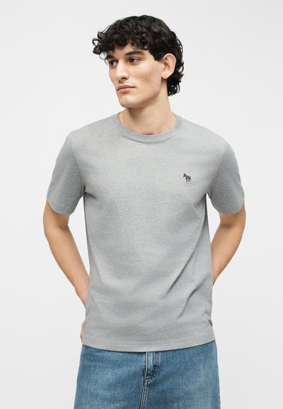 Νεαρός άνδρας με κοντά σγουρά μαύρα μαλλιά που φοράει απλό γκρι κοντομάνικο μπλουζάκι με μικρό κεντημένο λογότυπο ζέβρας στο στήθος.