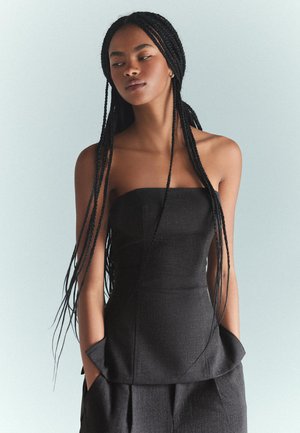 Črni top brez naramnic v peplum stilu, izdelan iz teksturirane tkanine, ki ima prilegajoč bodice in stranske žepe. Model pozira proti svetli poti.