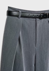 Pantalons plissés gris avec une ceinture en cuir noir passée dans les passants, sur fond blanc.