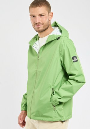 Homme portant une veste à capuche vert clair avec fermeture éclair et poches zippées, debout avec une main dans la poche devant un fond uni.