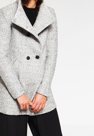 Femme portant un manteau gris texturé à double boutonnage avec de grands revers, des boutons noirs, et un pantalon noir, les mains jointes devant.