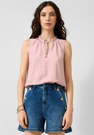 Frau trägt ärmellose hellrosa Bluse mit Rüschen am Kragen und hoch taillierte blaue Jeansshorts mit goldenen Knöpfen, steht vor einfarbigem Hintergrund.