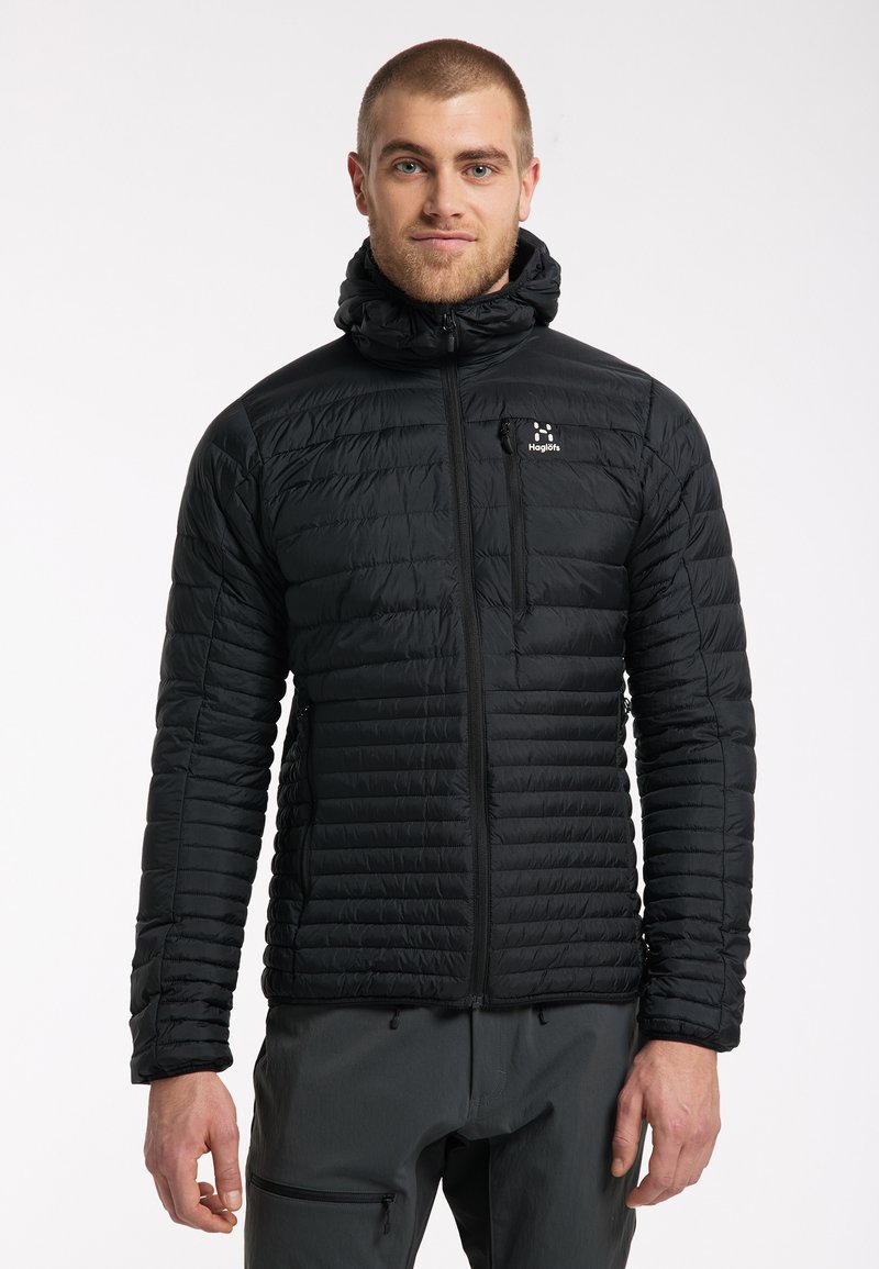 Chaqueta aislante negra con cierre de cremallera y capucha. Presenta textura acolchada, un bolsillo en el pecho y un logo sutil en el lado izquierdo.