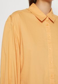 Chemise boutonnee orange clair avec un col, manches longues, texture lisse et boutons assortis sur le devant.