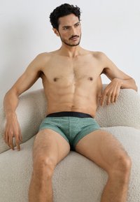 Zaļi rakstaini bokseršorti ar melnu elastīgu jostu, uz kuras ir logo teksts, tos valkā krekla nevalkājošs vīrietis, kas sēž uz teksturēta bēša dīvāna.