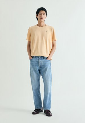 Hombre adulto con camiseta naranja pálida, vaqueros azul claro y zapatos marrón oscuro, de pie con las manos en los bolsillos frente a un fondo liso.