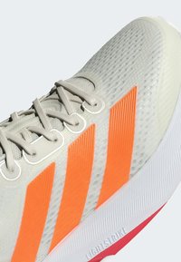 Zapatilla de running ligera con un upper texturizado en gris, tres franjas de color naranja brillante y una suela blanca con un acento rojo destacado.
