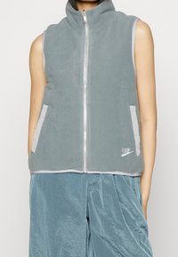 Grå fleeceväst med hög krage, framdragkedja och sidofickor. Kantad i ljusgrått, har en liten Nike-logotyp framtill.