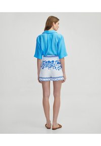Camicia blu a maniche corte con colletto; pantaloni corti bianchi con motivi floreali blu e orlo decorativo, abbinati a sandali marroni.