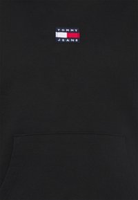 Sudadera negra con un pequeño logo bordado "TOMMY JEANS" en acentos rojos, blancos y azules. Incluye un bolsillo frontal. Textura suave.