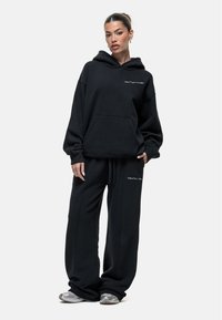 Zwarte hoodie en trainingsbroek set gemaakt van zacht materiaal. De hoodie heeft een voorzak, met een minimalistisch wit logo op de borst en de broek.