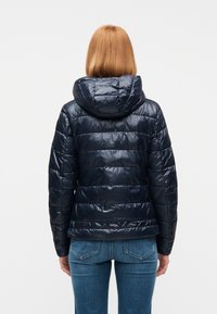 Γυναίκα με ίσια κόκκινα μαλλιά, που φοράει γυαλιστερό σκούρο μπλε μπουφάν με κουκούλα και μπλε τζιν, ορατή από πίσω.