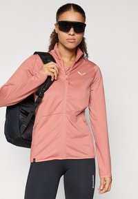 Helle rosa Zip-Jacke aus strukturiertem Stoff mit orangefarbenen Akzenten und zwei Seitentaschen. Modell trägt einen schwarzen Rucksack und Leggings.