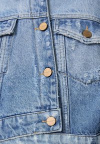 Chaqueta de mezclilla en color azul claro, con textura desgastada, botones de metal y dos bolsillos frontales con detalles de costura visibles.