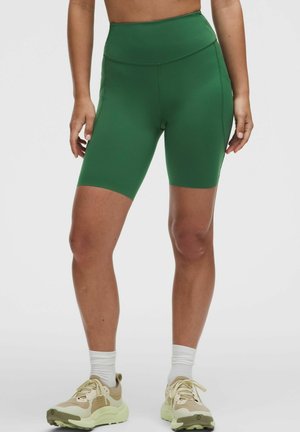 Femme portant un short de vélo vert taille haute, des chaussettes blanches et des baskets beige clair, debout devant un fond clair uni.