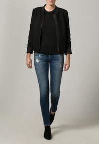 Chaqueta bomber negra con cremallera frontal y puños acanalados, combinada con jeans skinny azules desgastados. Botines negros completan el look.