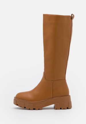 Tall brown leather boot with chunky lug sole and pull tab at back, shown from side on white background.