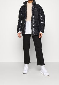 Giacca a vento nera con texture lucida, chiusura con zip e logo FILA. Sovrapposta a un maglione beige, abbinata a pantaloni neri e sneakers bianche.