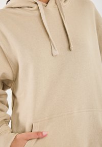 Sudadera beige hecha de tejido suave, con un bolsillo canguro, cordones ajustables y un corte relajado con puños de canalé.