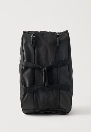 Musta suorakulmainen duffel-laukku, jossa on kaksi pystysuoraa vetoketjullista taskua ja kaksinkertaiset kantohihnat, jotka on sidottu keskeltä, pystyssä valkoisella pinnalla.