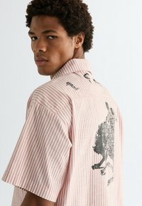 Πουκάμισο με κοντά μανίκια, ριγέ σε ροζ και λευκό, με γραφικό σχέδιο στην πλάτη. Γιακάς με κουμπιά, απαλό ύφασμα με άνετη εφαρμογή.