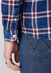 Modrá kostkovaná košile se červenými a bílými pruhy, s manžetou na knoflík a džíny z denimu s jemným detailem kapsy. Ruka spočívá na boku.