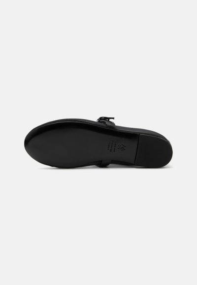 Chaussure noire à semelle plate vue par le dessous, avec un bout arrondi et une bride à la cheville avec boucle sur un fond gris uni.