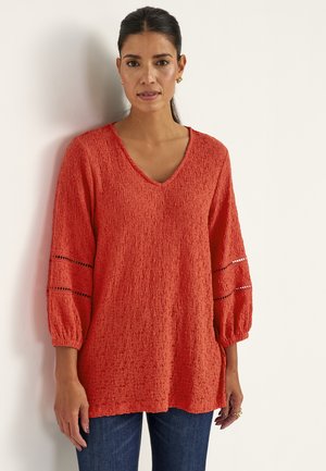 Kvinde iført rød langærmet top med tekstur og blonder på ærmerne samt blå jeans, stående foran en hvid baggrund.