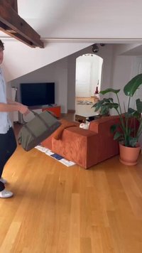 Olive green canvas duffel bag with dual handles, carried by a person in a room with wooden flooring, orange couch, and large potted plant.