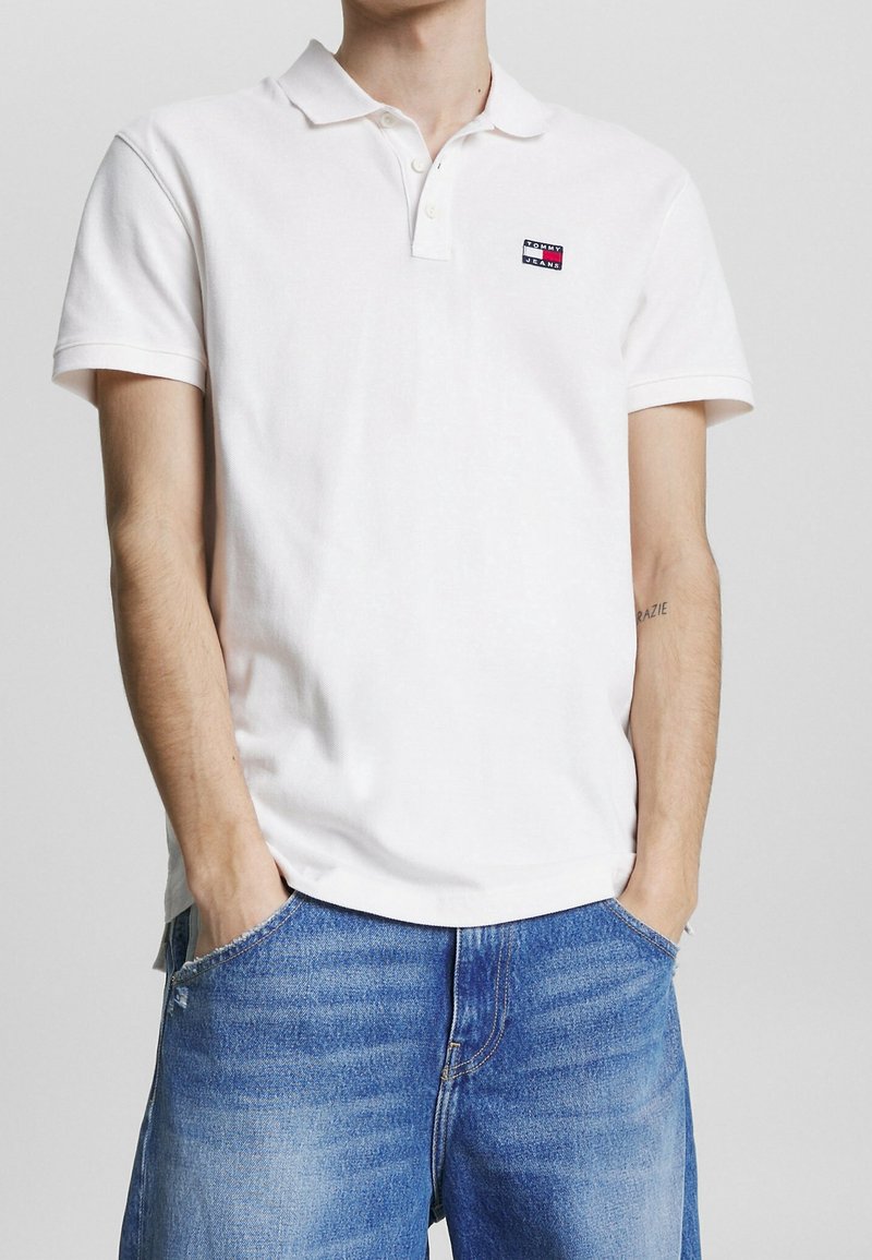Polo en coton blanc à manches courtes, avec un col et un petit logo rectangulaire rouge, bleu et blanc sur la poitrine gauche.