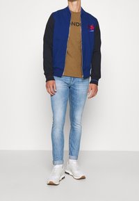 Marine bomberjakke med sorte ærmer, frontlynlås, blå accenter, brun t-shirt med sort tekst, lyseblå jeans, hvide sneakers.
