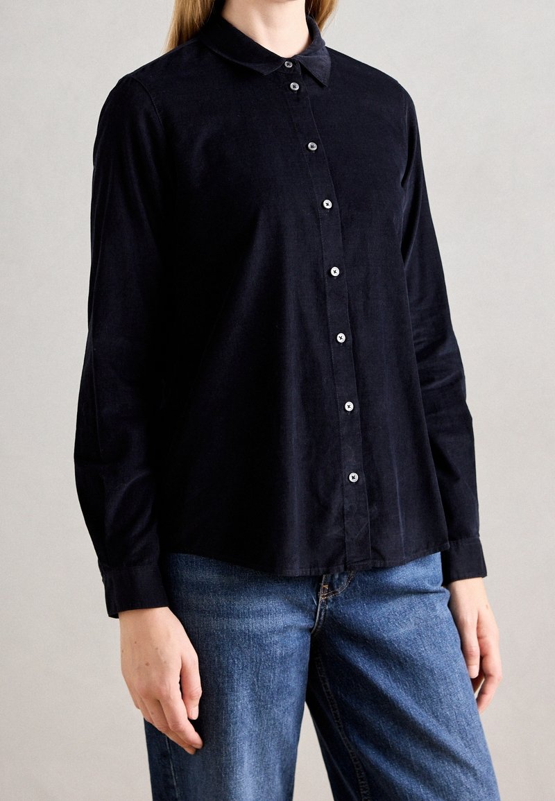 Personne portant une chemise noire à manches longues boutonnée avec un col et un jean bleu, debout devant un fond clair uni.