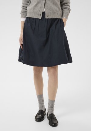 Frau trägt einen marineblauen knielangen Rock, eine graue, mit Knöpfen versehene Strickjacke, graue Socken und schwarze Loafers vor weißem Hintergrund.