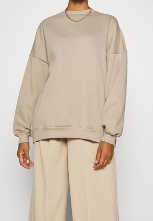 Persona con un suéter beige holgado con hombros caídos, pantalones beige de pierna ancha y un collar corto de cadena dorada sobre un fondo blanco.