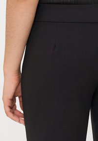 Pantalons noirs fuselés en tissu lisse, dotés d'une taille plate et d'une couture de taille discrète. Main reposant sur le côté.