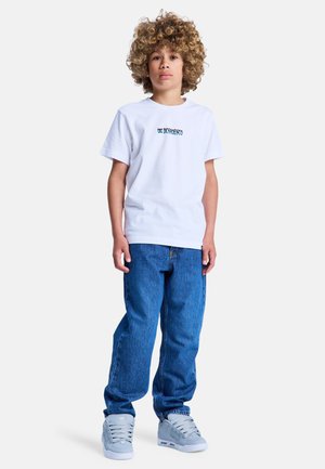Wit katoenen T-shirt met "DC Shoes"-logo, blauwe spijkerbroek en grijze sneakers. Kind staat met kort gekruld haar tegen een neutrale achtergrond.