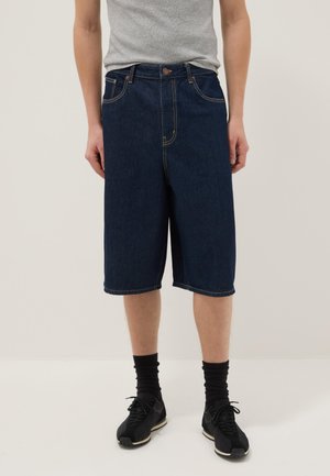 Persona che indossa pantaloncini in denim blu scuro fino al ginocchio, una canotta grigia, calzini neri e scarpe da ginnastica nere.