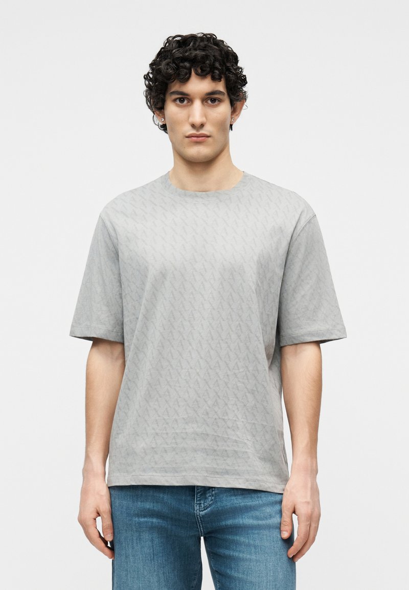 Νέος άνδρας με σγουρά μαύρα μαλλιά φοράει ανοιχτό γκρι μπλουζάκι με σχέδια και μπλε τζιν, στέκεται μπροστά σε απλή λευκή φόντο.