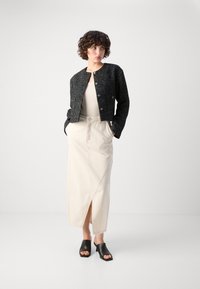 Giacca cropped nera con texture e bottoni argentati, top beige aderente, gonna midi color crema con spacco laterale e mules nere con tacco.