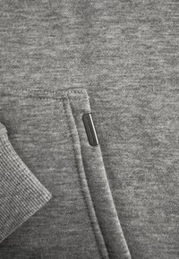 Close-up van grijze gebreide stof met mouwboord en zak, met een klein donker rechthoekig metalen merkje.