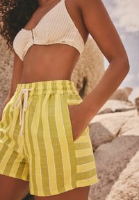 Gelb-weiße gestreifte Shorts mit einem elastischen Bund und Kordelzug, aus leichtem Stoff hergestellt. Verfügt über seitliche Taschen.