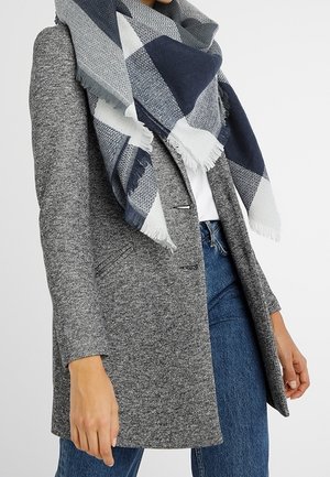 Femme portant un blazer gris texturé, une écharpe à carreaux bleus et blancs, un haut blanc et un jean bleu sur un fond uni.